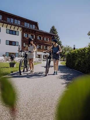 Two E-Bikers Walking Their Bikes in Front of the Hotel – Naturresort Puradies, Leogang, Salzburger Land Two e-bikers in front of the hotel, Naturresort Puradies in Leogang