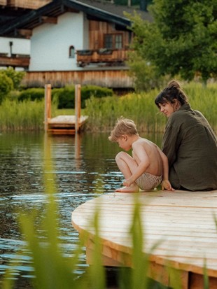 Naturbadeteich Heaven Spa – Mutter und Kind am Steg Mutter und Kind sitzen am Holzsteg des Naturbadeteichs im Heaven Spa, umgeben von Schilfgras, im Hintergrund die Chalets des PURADIES