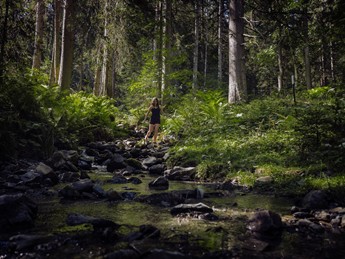 Wanderin balanciert über Steine im klaren Waldbach – Naturresort Puradies, Leogang, Salzburger Land Wanderin balanciert über Steine im Waldbach, Naturresort Puradies in Leogang
