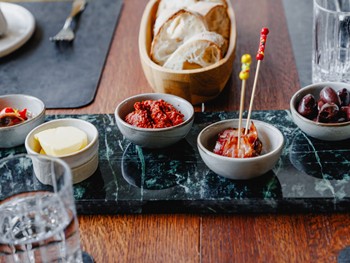 Tapas-Schälchen auf grünem Marmor im Restaurant Ess:enz – Naturresort Puradies Leogang Nahaufnahme von Tapas-Schälchen mit Butter, Tomatendip, Speckspieß und Oliven auf grünem Marmor sowie frisches Weißbrot in Holzschüssel im Restaurant Ess:enz, Puradies Leogang