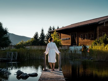 Person im Bademantel auf Steg am Naturbadeteich mit Saunahaus im Hintergrund bei Sonnenaufgang – Naturresort Puradies, Leogang, Salzburger Land Person im Bademantel auf Steg am Naturbadeteich bei Sonnenaufgang, Puradies Leogang