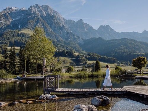 Person im Bademantel auf Holzsteg am Naturbadeteich vor Bergpanorama – Naturresort Puradies, Leogang, Salzburger Land Person im Bademantel auf Steg am Naturbadeteich, Puradies Leogang
