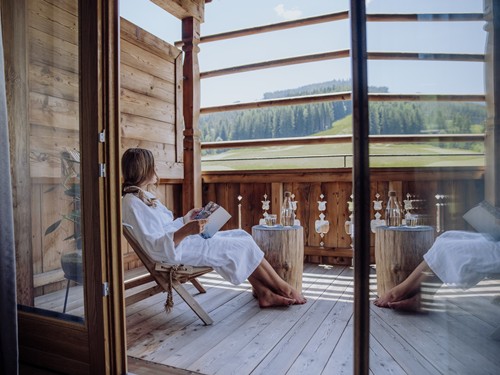 Person im Bademantel beim Lesen auf dem Balkon mit Blick auf grüne Landschaft – Naturresort Puradies, Leogang, Salzburger Land Person im Bademantel auf Balkon mit Landschaftsblick, Puradies Leogang