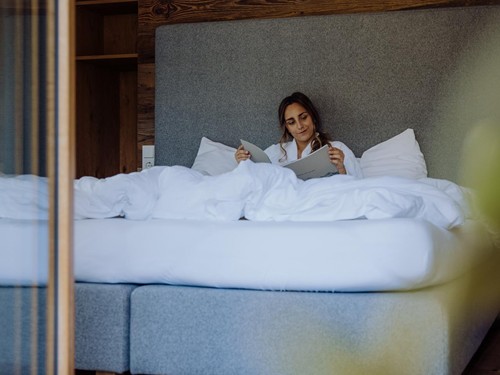 Person im Bademantel beim Lesen im Boxspringbett – Naturresort Puradies, Leogang, Salzburger Land Person im Bademantel liest im Bett, Puradies Leogang