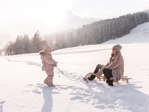Kind zieht Person auf Holzschlitten durch Winterlandschaft bei Naturresort Puradies Kind zieht Person auf Holzschlitten durch Winterlandschaft bei Naturresort Puradies