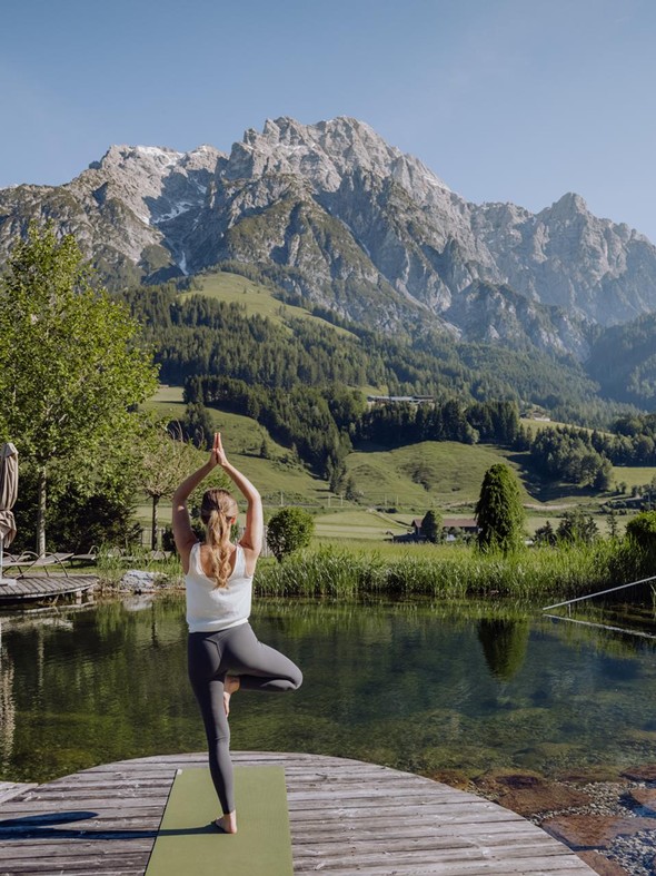 Yoga-Baumhaltung am Naturbadeteich mit Bergpanorama – Naturresort Puradies, Leogang, Salzburger Land Person in Yoga-Baumhaltung am Naturbadeteich, Naturresort Puradies in Leogang