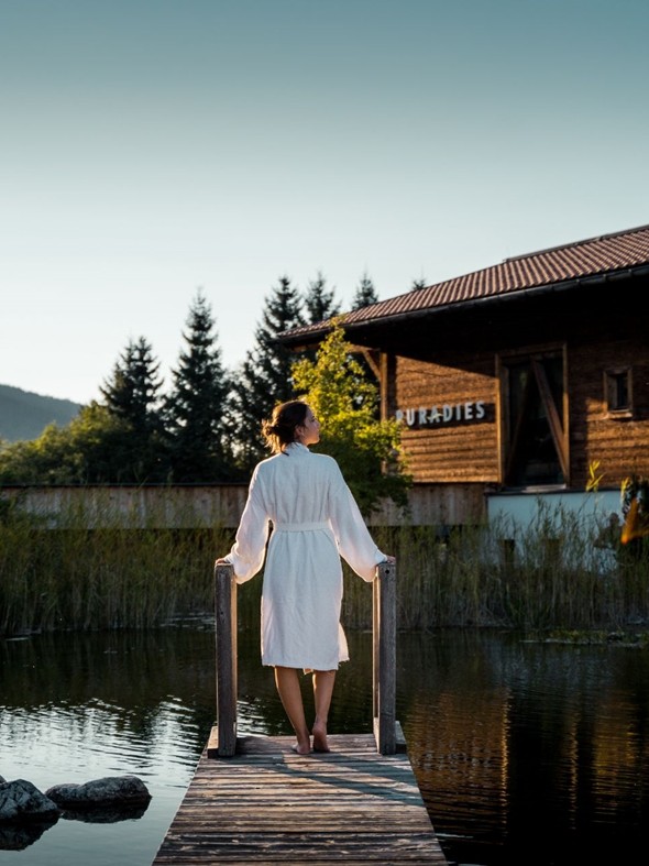 Person im Bademantel auf Steg am Naturbadeteich mit Saunahaus im Hintergrund bei Sonnenaufgang – Naturresort Puradies, Leogang, Salzburger Land Person im Bademantel auf Steg am Naturbadeteich bei Sonnenaufgang, Puradies Leogang